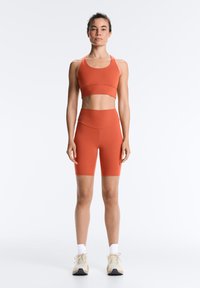 Woman standing facing forward wearing orange sports bra and matching high-waisted biker shorts with white sneakers against white background.