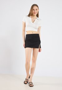 Vit polo crop top med korta ärmar, i kombination med en åtsittande svart mini kjol med en sidoflik. Modellen bär svarta klackskor med remmar.