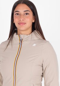 Jonge vrouw met lang donker haar in een beige jas met een meerkleuren gestreepte rits en een klein logo op de borst.