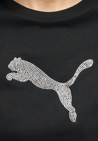 Λογότυπο πούμα που πηδάει, σχηματισμένο από μικρές στρογγυλές ασημένιες τρουκ, σε μαύρο ύφασμα, στο κέντρο του πάνω μέρους του στήθους ενός πο υκάμισου.