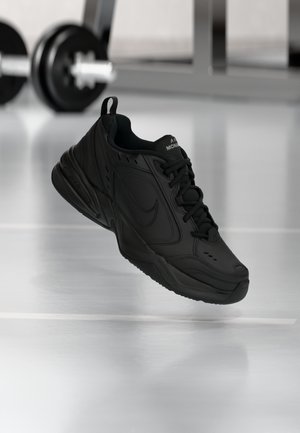 Baskets de sport noires avec lacets et logo swoosh, flottant au-dessus d’un sol de salle de sport réfléchissant avec des poids flous en arrière-plan.