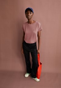 Rosa kurzärmliges T-Shirt mit umgeschlagenen Ärmeln, schwarze Hose, weiße und grüne Sneakers und eine rote Tragetasche, vor einem rosa Hintergrund.