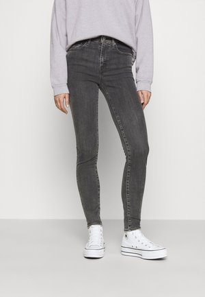 Person iført falmede sorte skinny jeans, lys grå sweatshirt og hvide højtopede sneakers, der står mod en ensfarvet baggrund.
