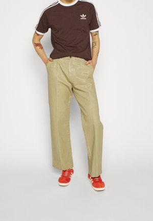 Maroon bomulds-T-shirt med hvide accenter og adidas-logo, parret med lysebeige brede bukser og røde ruskinds sneakers.