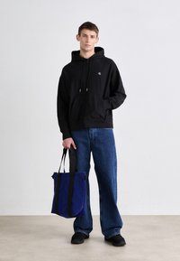 Čierna mikina s kapucňou a predným vreckom, kombinovaná s voľnými modrými džínsami a modrou taškou s čiernymi popruhmi. Neformálna obuv.