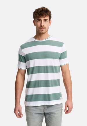 Młody mężczyzna stoi frontem do obserwatora, ubrany w białą koszulkę z szerokimi zielonymi poziomymi paskami oraz jasnoszare dżinsy.