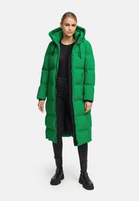 Groene pufferjas met een capuchon, voorzien van een ritssluiting, een gewatteerd patroon en een donkere voering, gecombineerd met zwarte jeans en stevige zwarte laarzen.