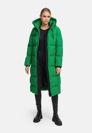 Groene pufferjas met een capuchon, voorzien van een ritssluiting, een gewatteerd patroon en een donkere voering, gecombineerd met zwarte jeans en stevige zwarte laarzen.