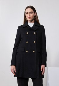 Manteau noir croisé en tissu texturé, doté de boutons dorés, de poches latérales et d'un col cranté.