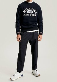 Mężczyzna noszący granatowy sweter z napisem "Hilfiger 1985 New York", ciemne wzorzyste spodnie, czarne skarpetki i białe trampki na neutralnym tle.