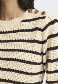 Beżowy sweter z dzianiny w ciemno-brązowe poziome paski, z trzema okrągłymi mosiężnymi guzikami na ramieniu, noszony przez osobę z długimi ciemnymi włosami.