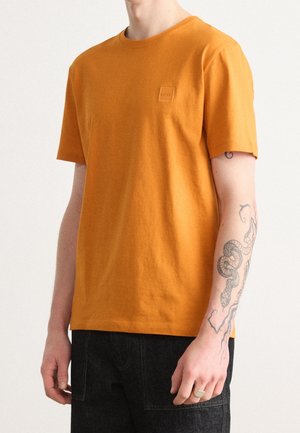Uomo che indossa una maglietta arancione semplice a maniche corte e pantaloni neri, mostrando un avambraccio destro tatuato con un disegno di serpente.
