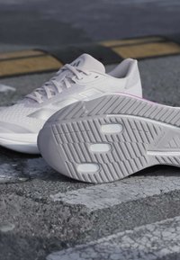 Weiße Sportschuhe auf Asphalt, ein Schuh steht aufrecht und der andere zeigt die strukturierte Sohle mit Grifffeldern.