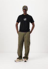 Tricou negru cu logo, pantaloni cargo verzi oliv și pantofi sport bej. Include, de asemenea, o șapcă cu un model camuflaj. Fundal simplu.