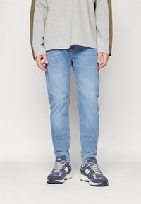 Jeans de mezclilla azul claro de corte ajustado, combinados con una camiseta de manga larga gris con detalles en verde oliva. Se llevan con zapatillas deportivas en color azul marino.