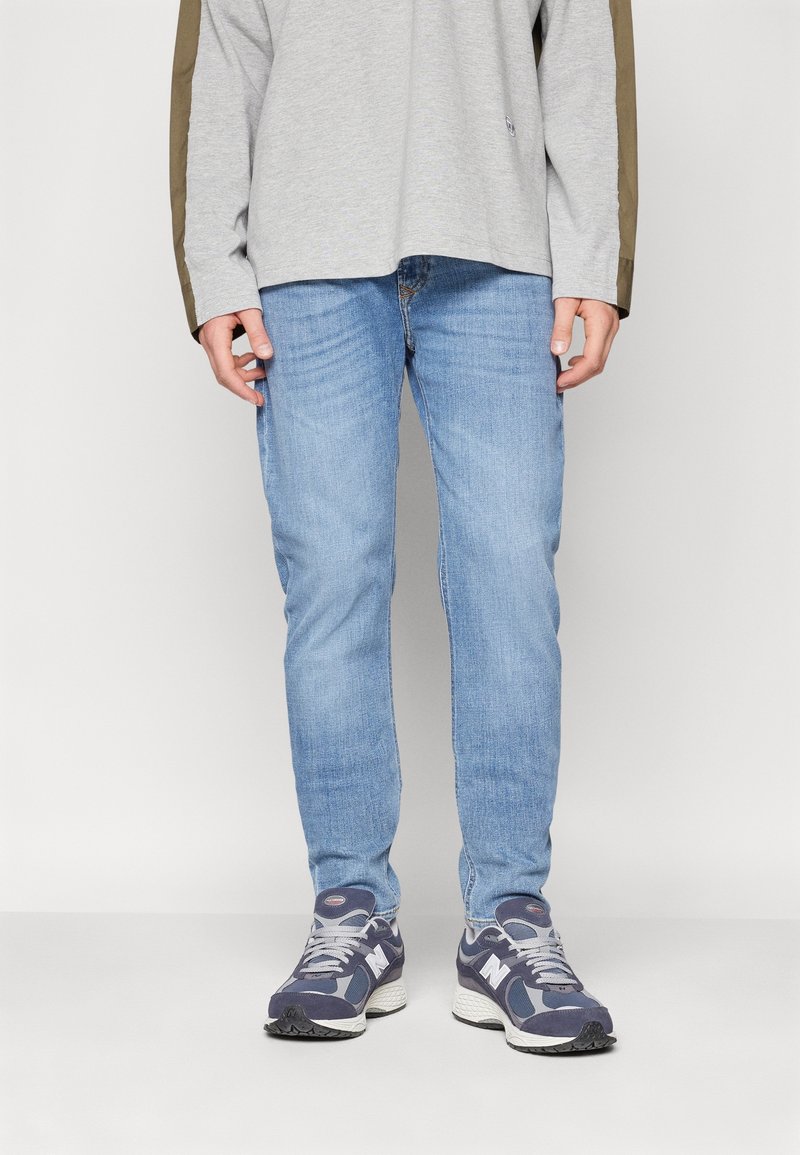 Jeans de mezclilla azul claro de corte ajustado, combinados con una camiseta de manga larga gris con detalles en verde oliva. Se llevan con zapatillas deportivas en color azul marino.