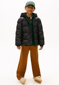 Chaqueta acolchada negra con capucha, camiseta polo verde, pantalones de pierna ancha marrones y zapatillas beige, con fondo blanco.