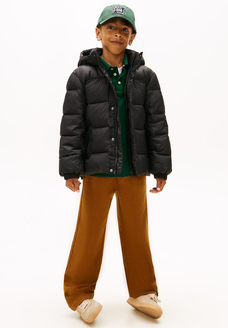 Chaqueta acolchada negra con capucha, camiseta polo verde, pantalones de pierna ancha marrones y zapatillas beige, con fondo blanco.