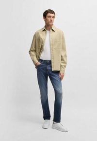 Beige knäppskjorta över en vit t-shirt, tillsammans med blå denimjeans och vita sneakers; har två bröstfickor och en avslappnad passform.