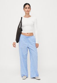 Top cropped de mangas longas brancas combinado com calças largas listradas em azul e branco. Modelo segura uma mala tote castanha-escura. Os sapatos são sapatilhas de cor clara.