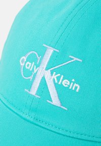 Gaiši zilā vāciņš, kas izgatavots no kokvilnas, ar balto izšūto "Calvin Klein" logo. Strukturā ir iekļauta izliekta malām un teksturēts audums.