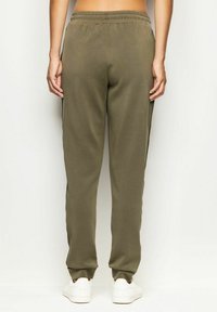 Osoba ubrana w oliwkowe spodnie dresowe z czarnymi bocznymi paskami i białe sneakersy, stojąca tyłem na białym tle.