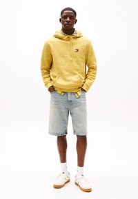 Felpa gialla con tasca frontale a canguro, pantaloni corti in denim e sneakers bianche. Accentuata da un dettaglio a scacchi sul colletto della felpa.