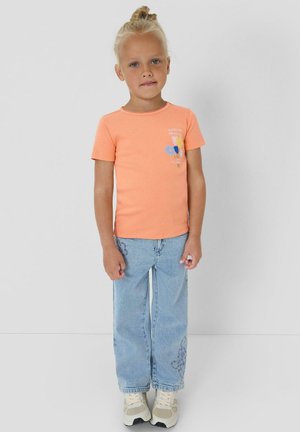 Junges Mädchen mit blondem Haar zu einem Dutt gebunden, trägt ein orangefarbenes kurzärmeliges Shirt mit Blumenmuster, hellblaue Jeans und weiße Sportschuhe.