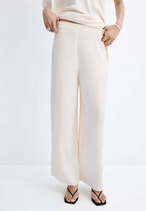 Mujer con pantalones crema de tiro alto y pernera ancha, con un top a juego, ajustándose la cintura y llevando sandalias negras tipo tanga.