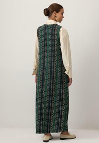 Rochie maxi cu un model geometric verde, negru și auriu; fără mâneci, cu guler înalt; purtată peste o cămașă deschisă la culoare.