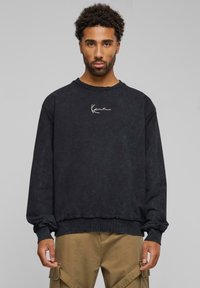 Zwarte, crewneck sweatshirt met een versleten textuur en zoom. Voorzien van een eenvoudig wit logo op de borst. Gecombineerd met tan cargobroeken.