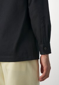 Black ribbed long-sleeve shirt with buttoned cuffs, featuring a straight hem and relaxed fit, paired with light yellow trousers.