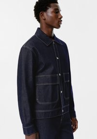 Giacca in denim blu scuro con chiusura a bottoni, due ampie tasche anteriori e cuciture gialle a contrasto. Dotata di colletto e vestibilità morbida.