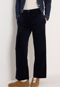 Navy Baumwollcordhosen mit weitem Bein und vorderen Taschen, kombiniert mit beigefarbenen Sneakers. Der Stoff hat eine gerippte Textur und eine weiche Optik.