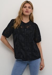 Blusa nera trasparente con motivi floreali, frontale arricciato, maniche corte e chiusura a bottoni, abbinata a jeans azzurri.