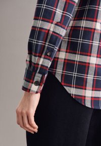 Checkered flannel shirt featuring navy, red, and white plaid design. Button cuffs and rounded hem, showcasing soft fabric texture.
