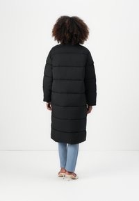 Black long puffer coat with horizontal quilted panels, featuring a high collar and long sleeves, worn over light blue pants and heels.