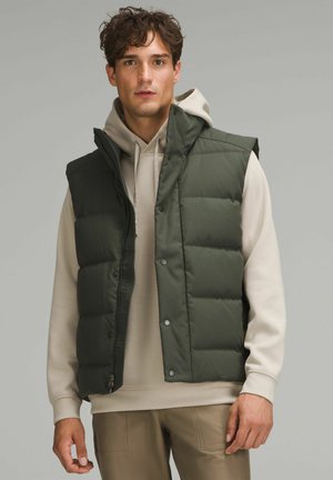 Polstret grøn vest med høj krave, trykknapper og lynlås. Båret over en beige hættetrøje. Beklædningen er glat med en matte overflade.