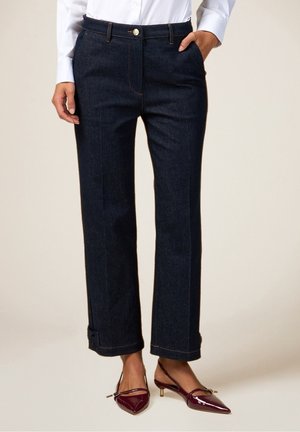 Nainen, joka pukeutuu tummansinisiin suoriin farkkuihin, joiden lahkeet on käännetty, ja viininpunaisiin korkeakorkoisiin slingback-kenkiin, käsi taskussa.