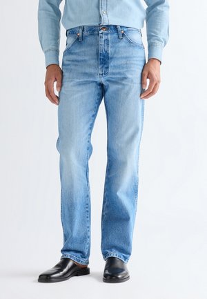 Mężczyzna ubrany w jasnoniebieskie jeansy o prostych nogawkach, niebieską koszulę na guziki i czarne wsuwane buty stojący na białym tle.