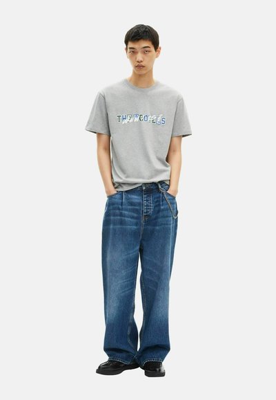 Gray cotton t-shirt with graphic text "THE WOLVES" in blue, paired with loose-fitting blue denim jeans and black shoes.