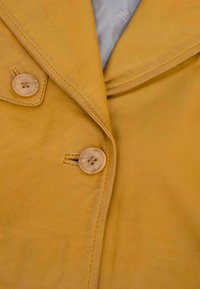 Senfgelbe Lederjacke mit Reverskragen, zwei Knöpfen mit holzartiger Oberfläche und glatter, leicht strukturierter Oberfläche.