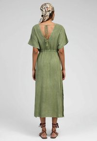 Robe midi vert olive avec des manches flottantes courtes, dos ouvert noué au cou, taille élastique et fentes sur les côtés, associée à des sandales à lanières brunes.