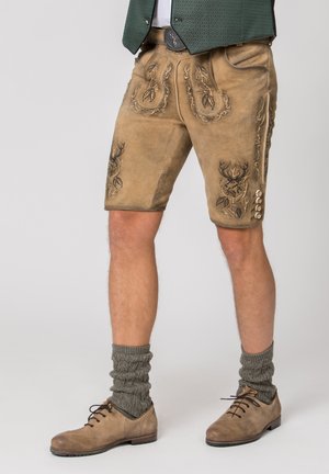 Mann trägt traditionelle braune Lederhosen-Shorts mit besticktem Hirsch, graue gestrickte Socken und braune Lederschuhe.