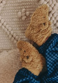 Rjave puhasti copate z rahlo teksturirano površino, oblikovane kot medvedje tace, nošene s temno modrimi karirastimi hlačami, na svetlem vzorčastem ozadju.