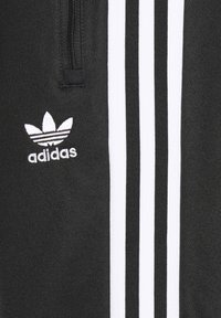 Svarte treningsbukser med hvit tre-stripesdesign, med en glidelåslomme og brodert Adidas-logo på venstre lår.