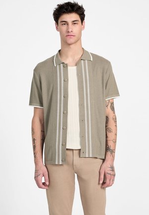Camisa de manga corta, color verde oliva, con rayas blancas, con botones en la parte delantera y cuello en punta. Usada sobre una camiseta de tirantes blanca con acanalado.