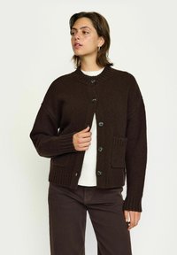 Hnědý pletený cardigan s kulatým výstřihem, předními knoflíky, žebrovanými manžetami a dvěma předními kapsami. Tkanina vypadá jemně a texturovaně.
