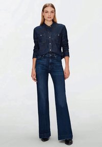 Woman standing wearing dark blue button-up denim shirt and high-waisted wide-leg dark blue jeans with black shoes.