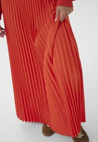 Persona, kas valkā spilgti oranžu plisētu svārku, tur audumu ar vienu roku, apakšā redzamas brūnas kedas.